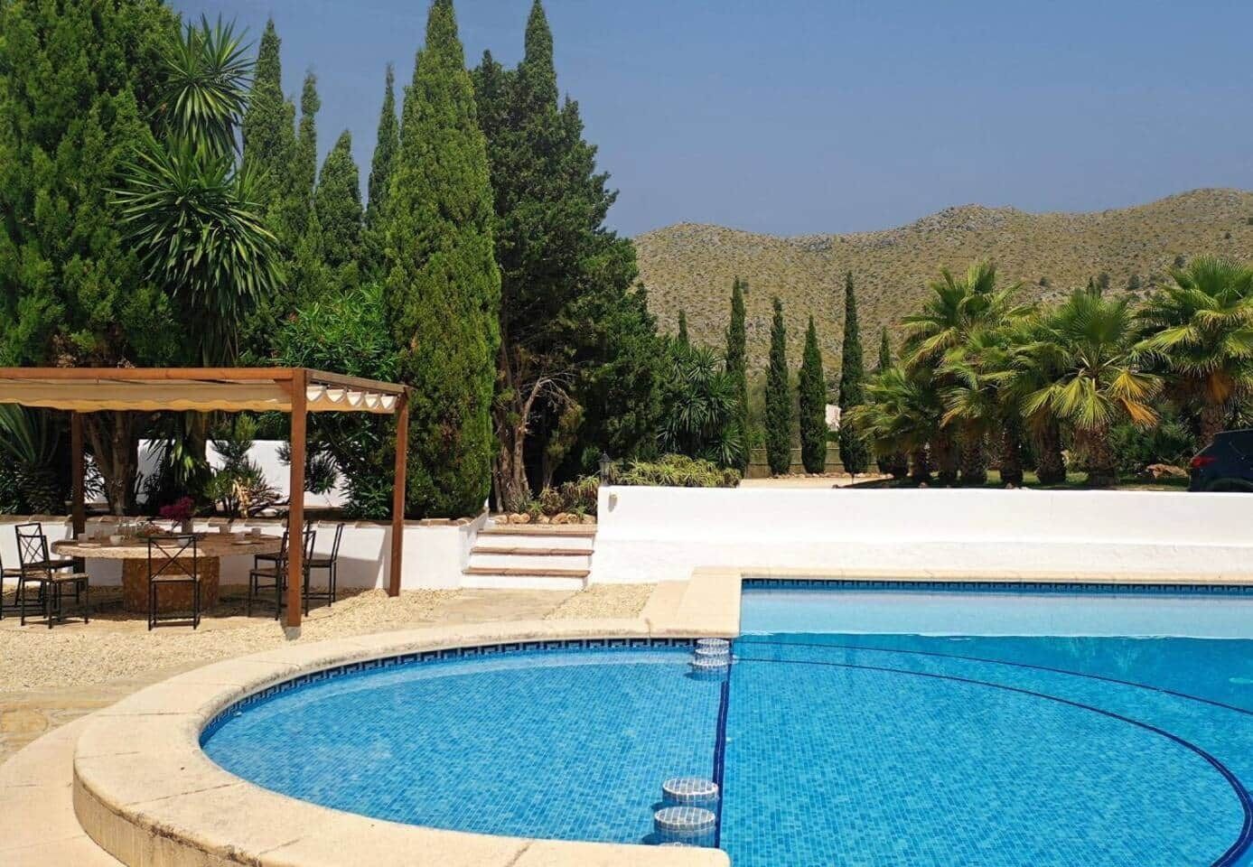 Une piscine agrémentée d’une pergola et de chaises, entourée de cyprès et de palmiers, avec une montagne en toile de fond.