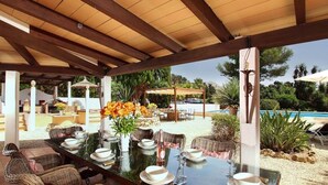 An outdoor dining area with a long table set for six, surrounded by wicker chairs, under a wooden pergola with a view of a swimming pool and greenery. - Villa Jacobus (Pollença)