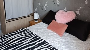 Makuuhuone, jossa on seepramustakuosinen päiväpeite, valkoinen verho, peili ja kukkakuviotapetti.