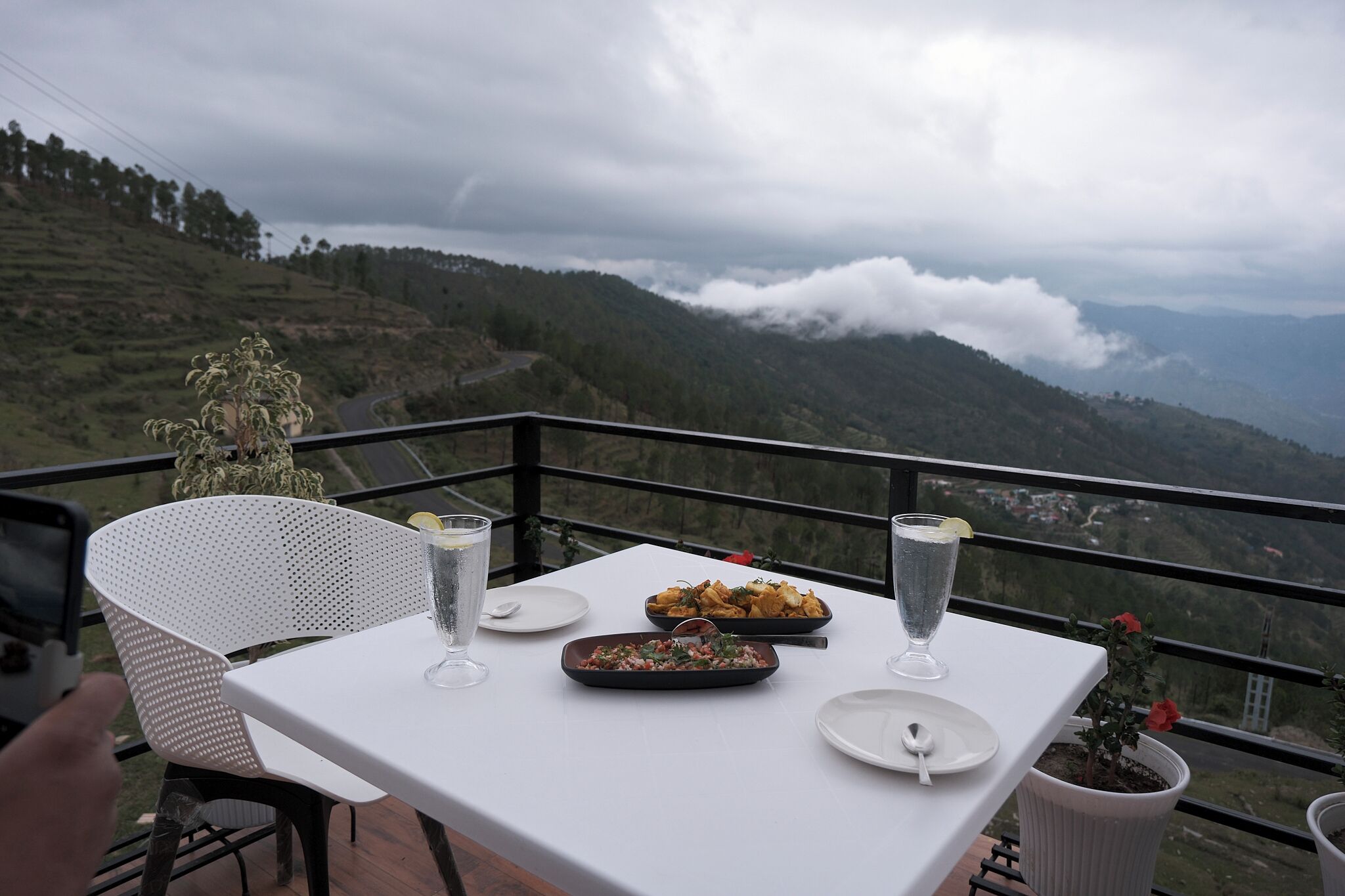 A table with dishes and drinks, a potted plant, and a view of a mountainous landscape.