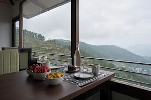 A dining table with bowls of fruit, a bottle, and a slice of cake, set against a view of a mountainous landscape. - Ivaara Resort (Pauri)