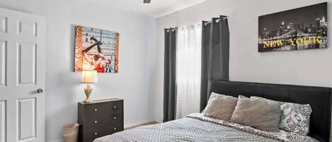 Een slaapkamer met een bed, een plafondventilator, een nachtkastje, een lamp, een poster en een raam met gordijnen.