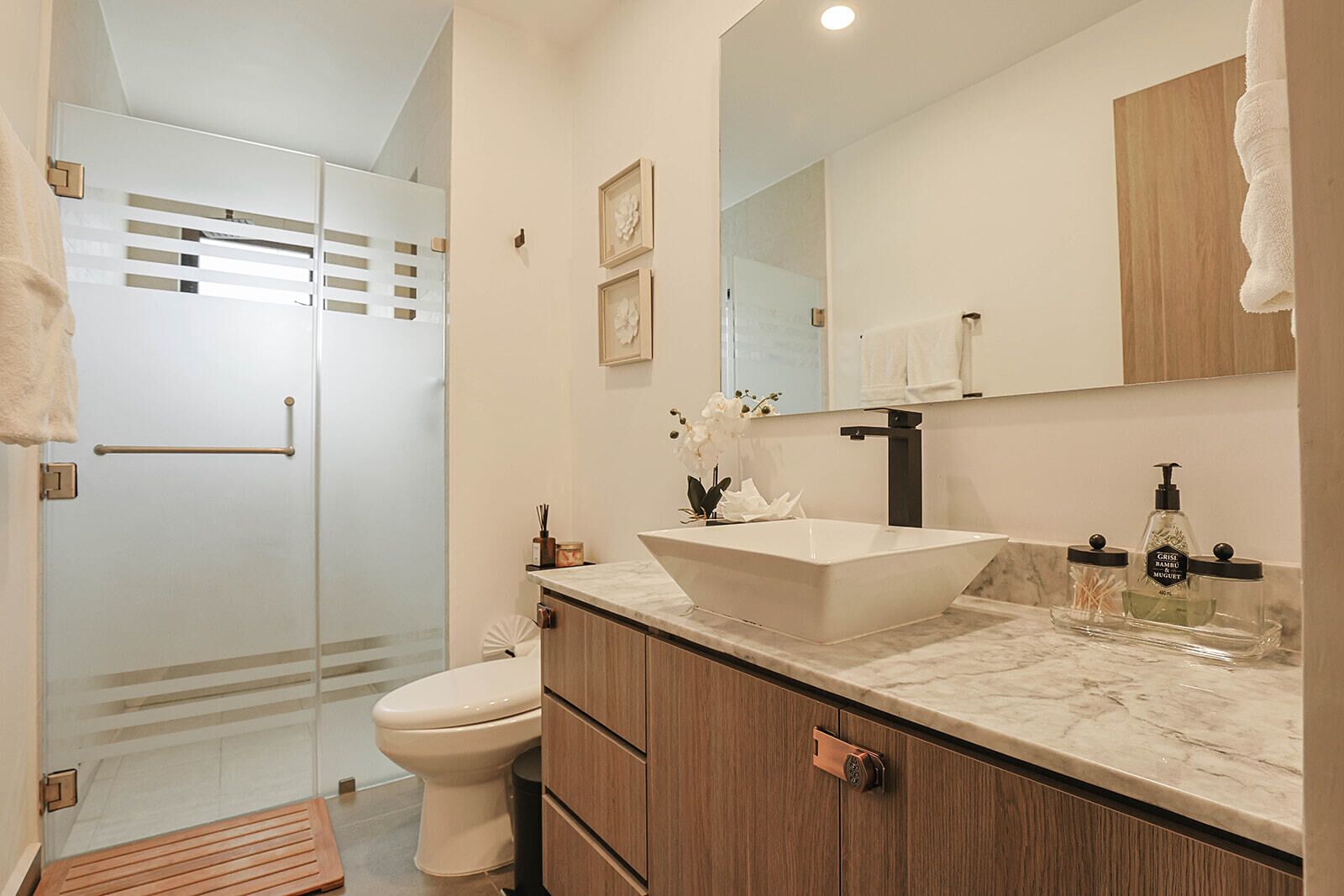Un baño moderno con cabina de ducha de vidrio, inodoro blanco, un lavabo de diseño sobre pedestal, armarios de madera y espejo sobre el lavabo.
