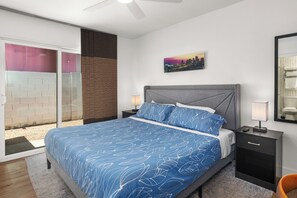 Ein Schlafzimmer mit einem Bett, einem Nachttisch, einer Lampe, einem Stadtbild als Gemälde und einer Schiebetür.