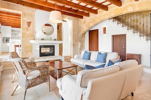 A living room with a fireplace, sofa, wicker chair, and a staircase. - Villa Alaior with private pool - My Rental Homes (Alaior)
