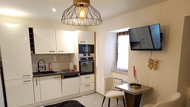 A modern kitchen with white cabinets, a black countertop, and a built-in oven. There is a dining area with a table and two chairs, and a TV mounted on the wall. - Charming studio in the heart of the mountains (Burzet)