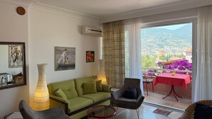 A living room with a green sofa, a glass coffee table, a chair, and a table with a red tablecloth. There are paintings on the wall and a view of a balcony with purple flowers. - Duplex House on the Castle Road with a Sea view (Alanya)