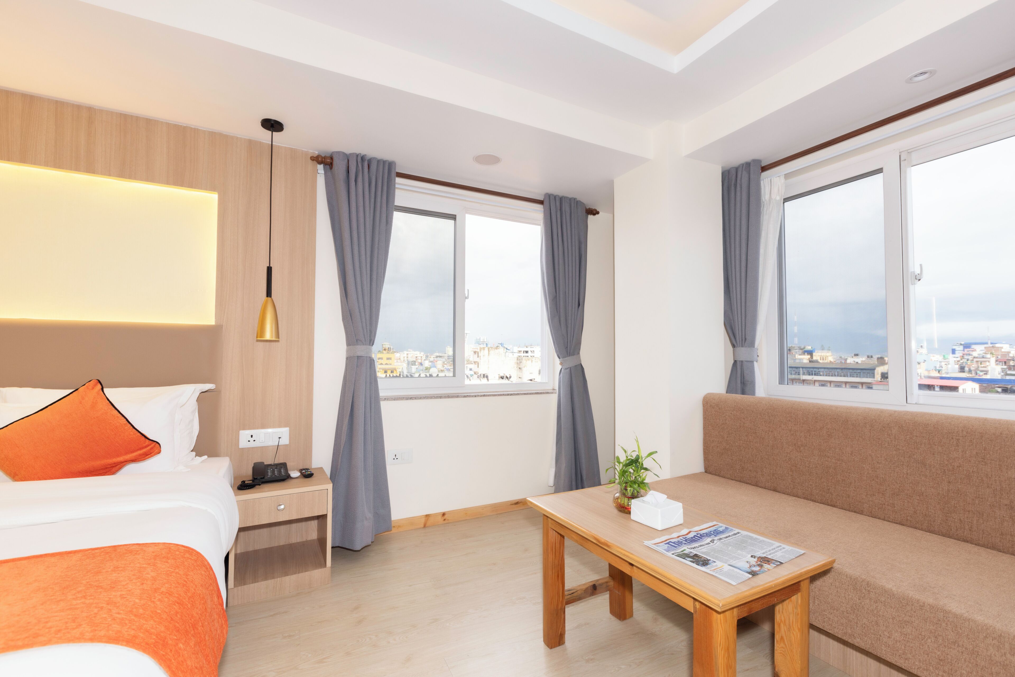 A hotel room with a bed, bedside table, lamp, curtains, a sofa, a small table with a plant, and a window with a city view.