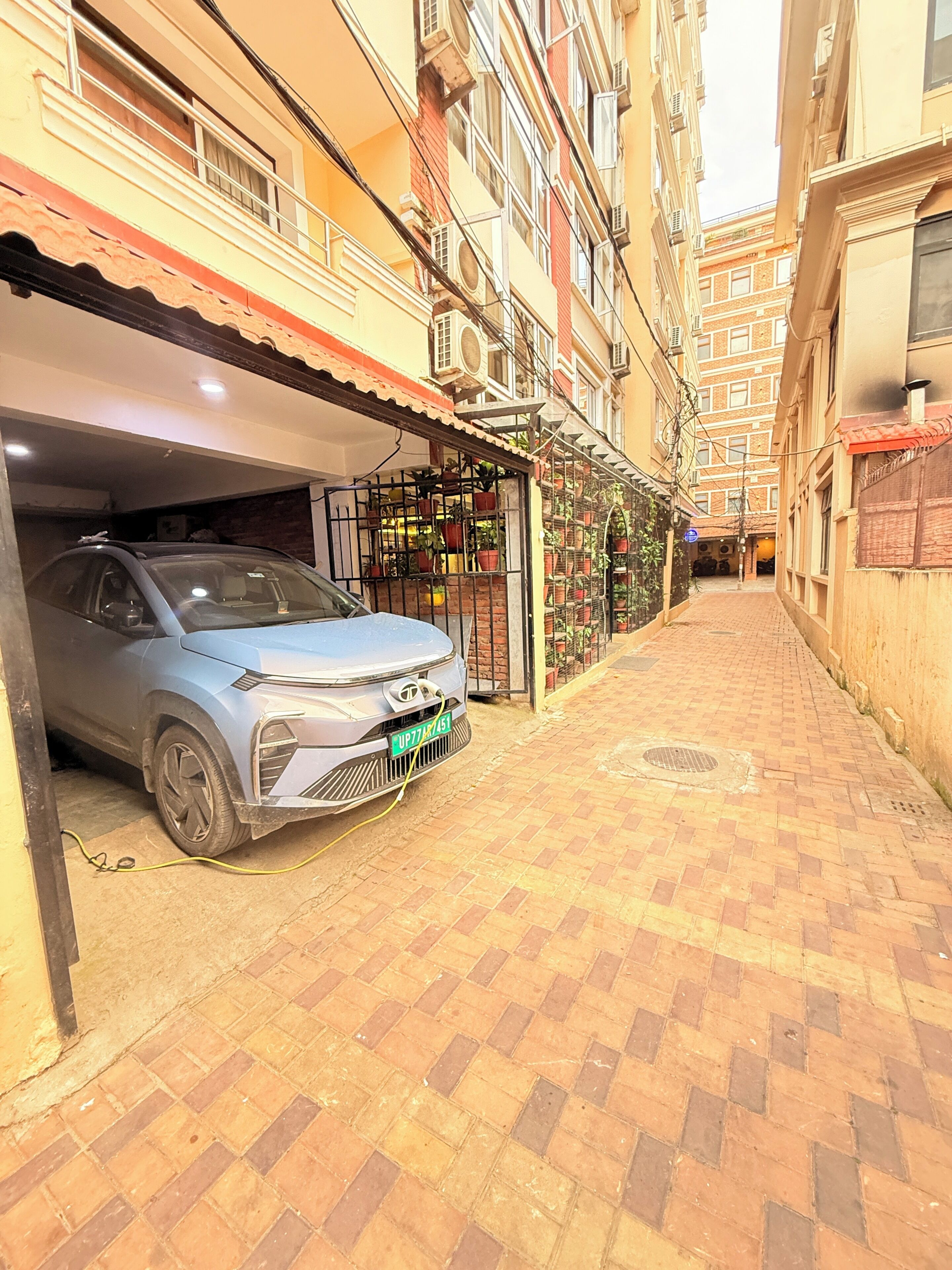 Un callejón estrecho con un coche estacionado, una entrada con rejas y plantas en macetas, y edificios de varios pisos con balcones.