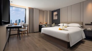 Une chambre d’hôtel moderne, dotée d’un grand lit, d’un bureau avec une chaise, d’une petite table ornée d’un vase de fleurs et offrant une vue sur la ville grâce à une grande fenêtre.