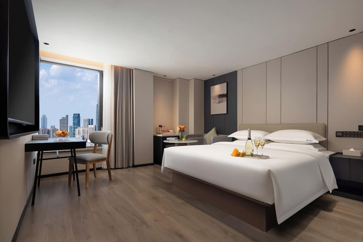 Een moderne hotelkamer met een groot bed, een bureau met stoel, een klein tafeltje met een bloemenvaas, en uitzicht op de stad door een groot raam.