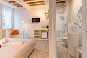 A hotel room with a bed, a small table, a chair, a TV, a shower, and a bathroom.