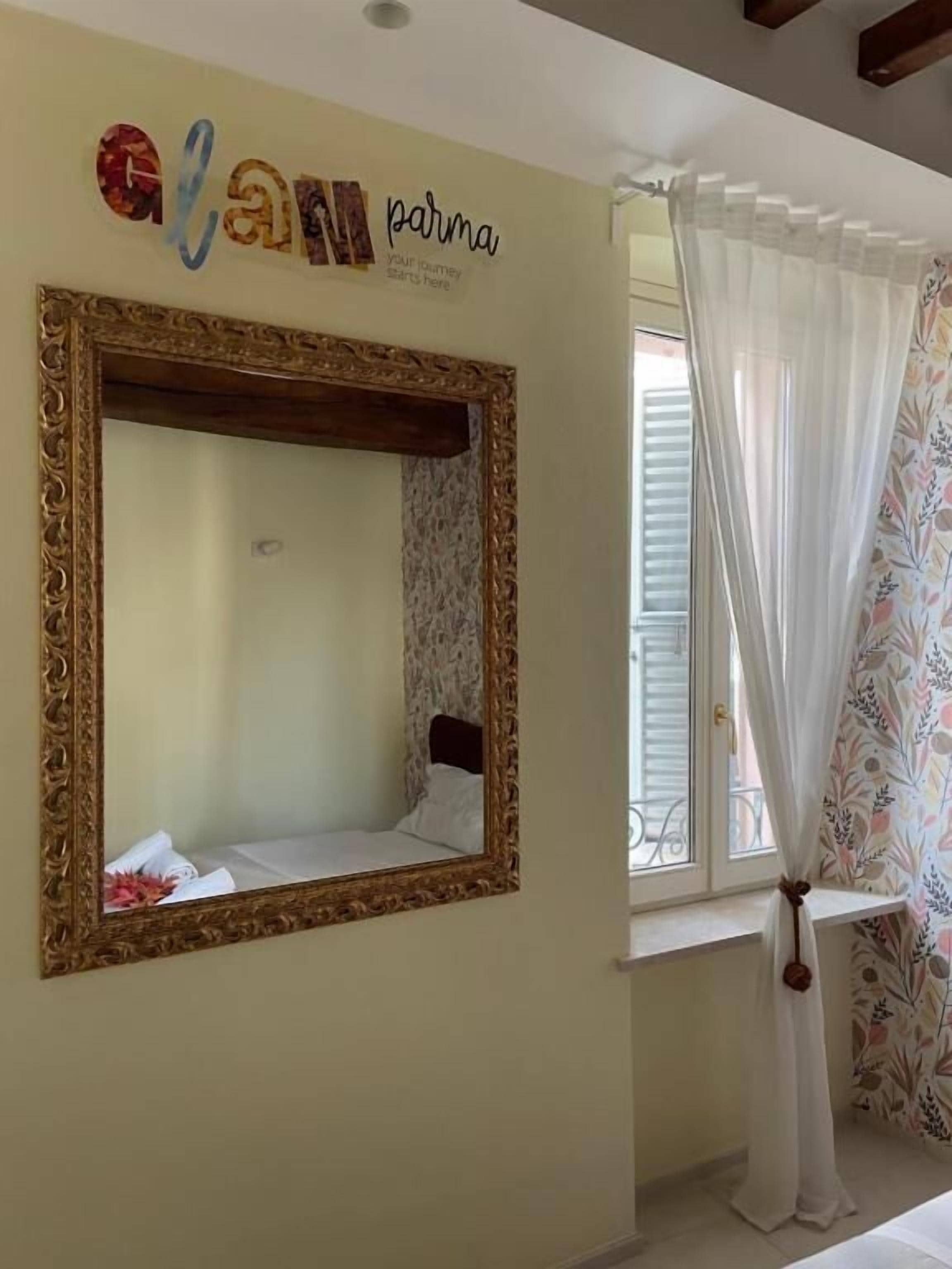 A room with a large ornate mirror, a bed with white bedding, a window with a floral curtain, and a wall with a decorative sign.