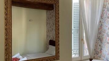 A room with a large ornate mirror, a bed with white bedding, a window with a floral curtain, and a wall with a decorative sign.