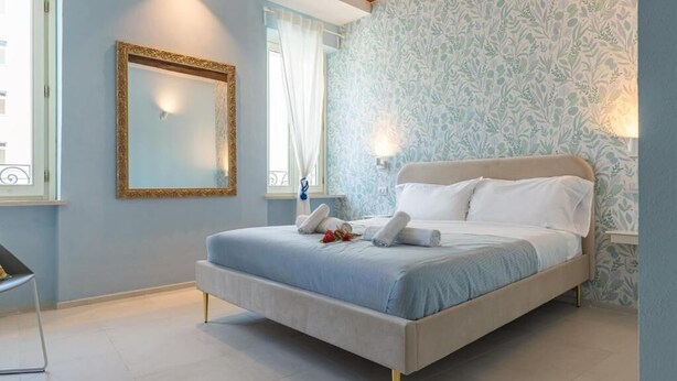 A bedroom with a bed, a mirror, a window with curtains, and a wall with a floral pattern. - GLAM PARMA (Parma)