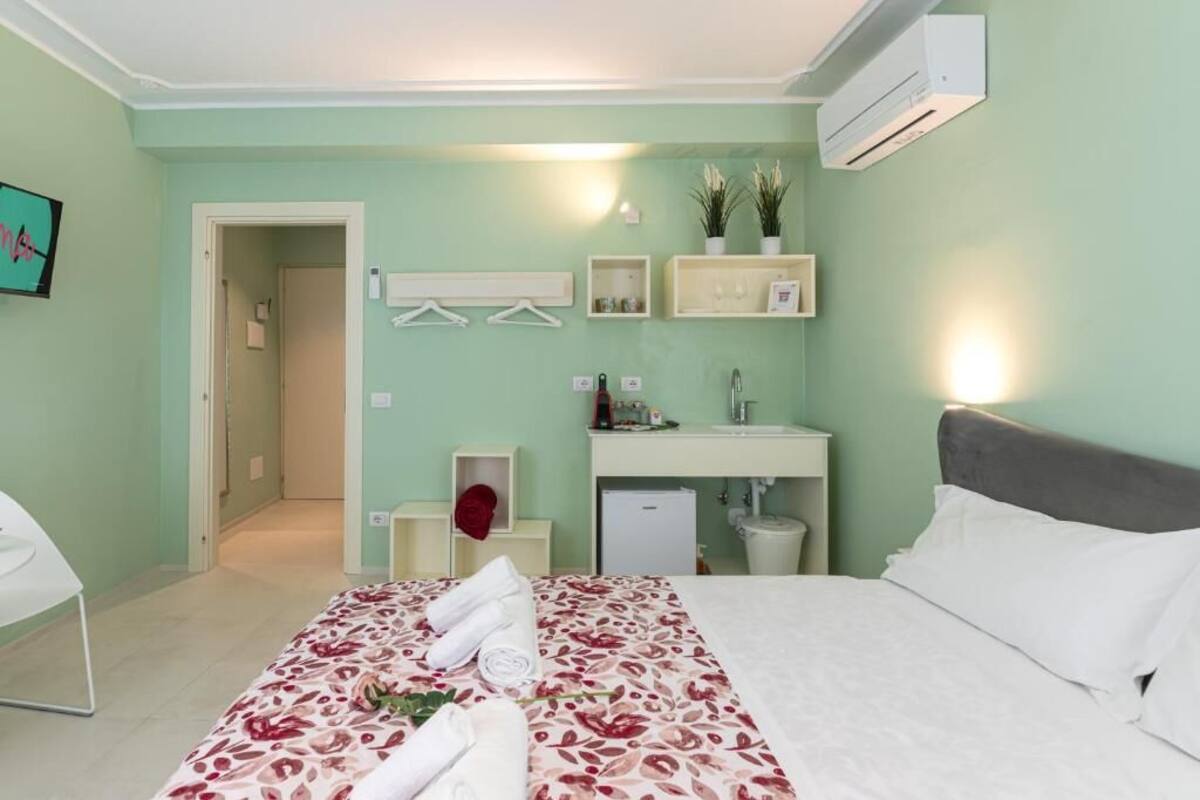 A bedroom with a bed, a bedside table, a nightstand, a wall-mounted air conditioner, a bathroom with a sink and mirror, and a hallway leading to another room.