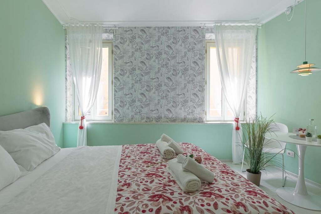 A hotel room with a bed, two windows with curtains, a bedside table with a lamp, and a small potted plant.
