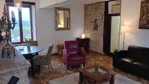 A living room with a stone wall, a dining table with chairs, a red armchair, a black leather sofa, and a chandelier. - Charming gîte with panoramic views over the hills of the Morvan (James)