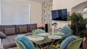 En moderne stue med en sofa, grønne lænestole, et glasbord, et fjernsyn og en plante.