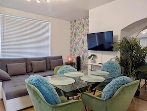Ein modernes Wohnzimmer mit einer Couch, grünen Sesseln, einem Glastisch, einem Fernseher und einer Pflanze.