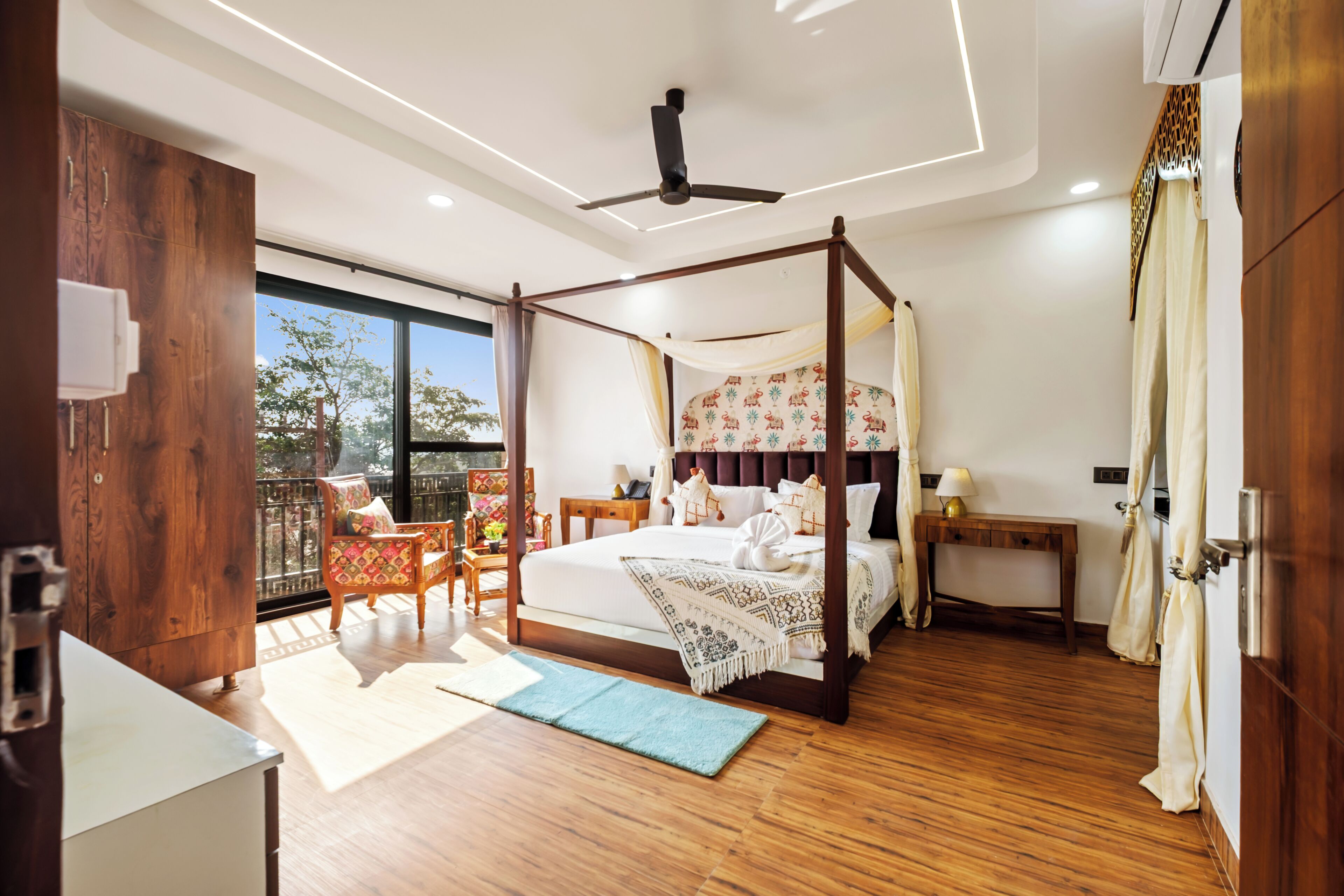 A bedroom with a four-poster bed, wooden flooring, a balcony with chairs, and a view of the outdoors.