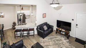 Uma sala de estar moderna com um sofá de couro preto, uma televisão e uma cozinha ao fundo.