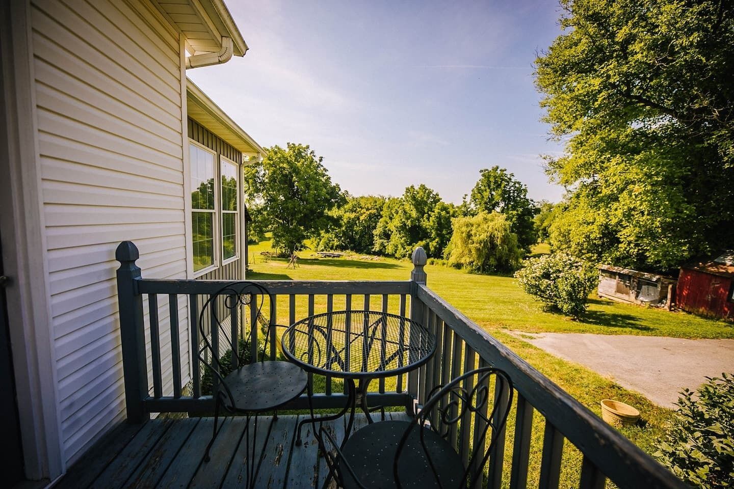 Une véranda avec une table et des chaises, une maison aux murs recouverts de bardage blanc et une pelouse verdoyante parsemée d’arbres.