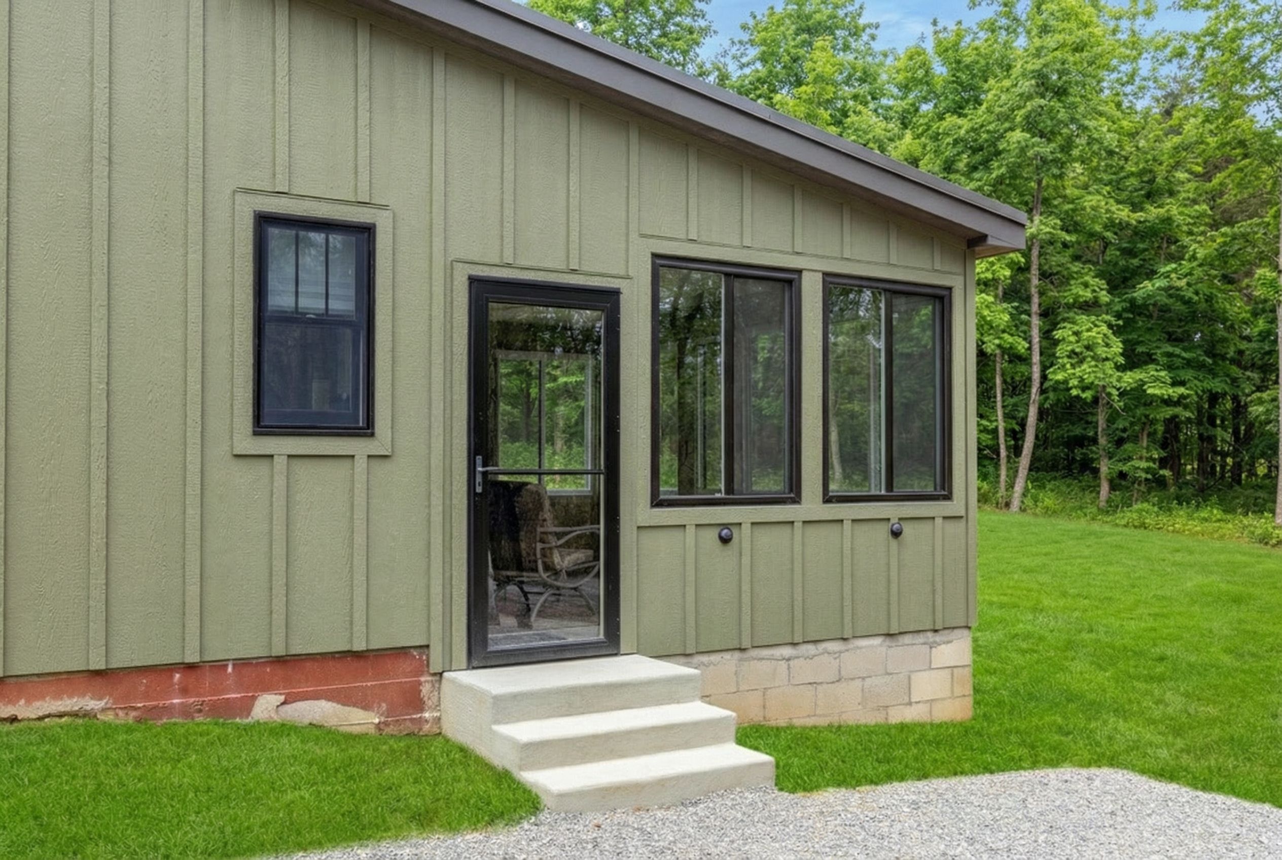 A single-story building with a green exterior, a front door, and a set of steps leading to a gravel driveway.