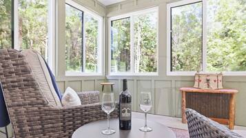 Una silla de mimbre, una mesa, una botella de vino y dos copas de vino en una habitación con ventanas amplias.