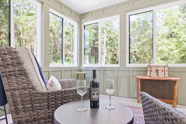 A wicker chair, a table, a bottle of wine, and two wine glasses in a room with large windows.