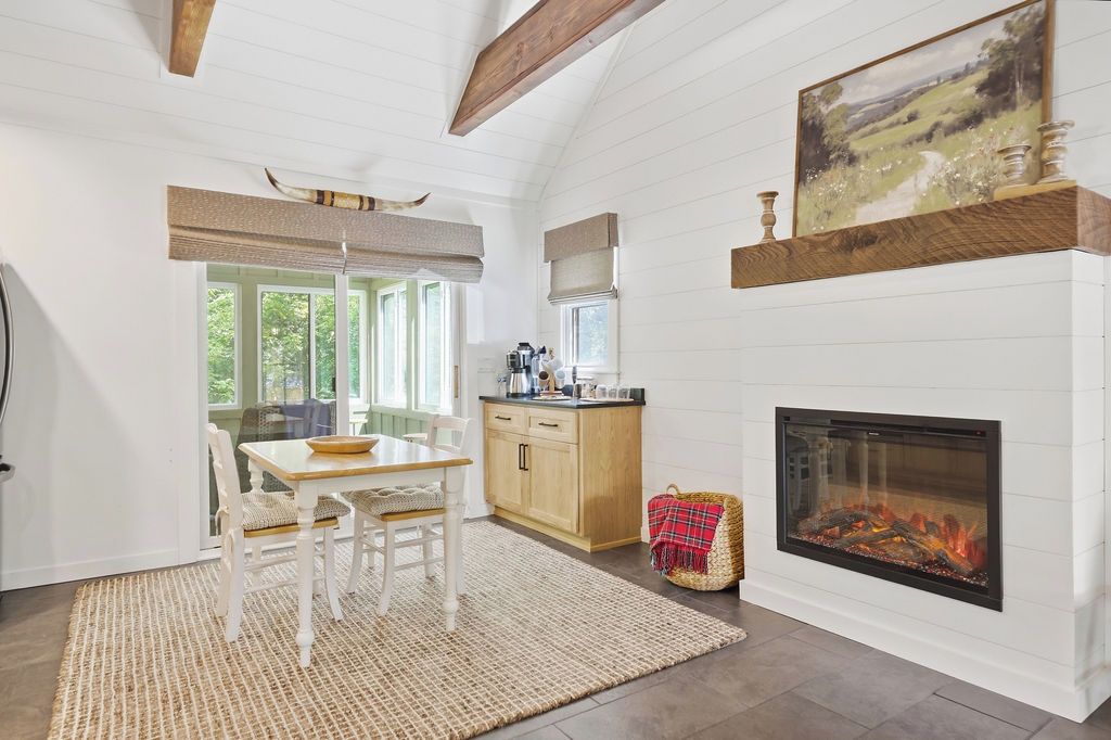 A room with a fireplace, a dining table with chairs, a kitchen area, and a window with a view of greenery.