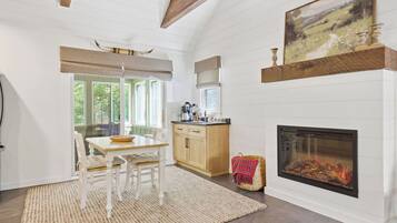 A room with a fireplace, a dining table with chairs, a kitchen area, and a window with a view of greenery.