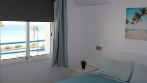 Un dormitorio con una cama, una ventana con vistas a la playa, una mesita de noche pequeña con una planta y un equipo de aire acondicionado en la pared.