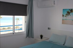 Ein Schlafzimmer mit Bett, Fenster mit Meerblick, einem kleinen Nachttisch mit Pflanze und einer Klimaanlage an der Wand.