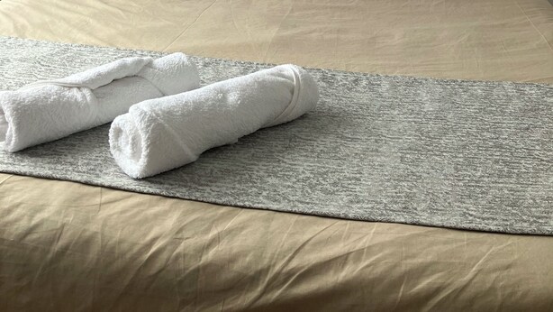 A neatly made bed with two pillows, a grey textured blanket, and two rolled towels. - Radama Hotel & Studios (Antananarivo)