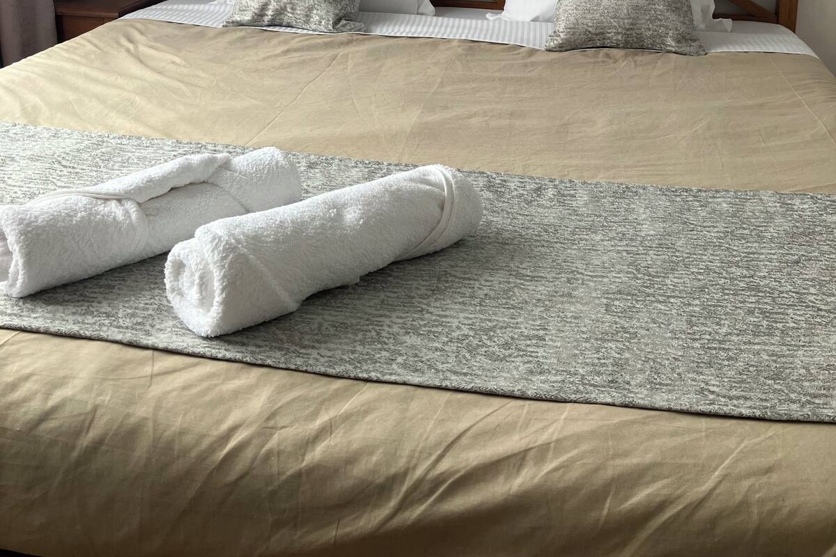A neatly made bed with two pillows, a grey textured blanket, and two rolled towels.