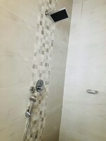 Zona de ducha con pared de azulejos, cabezal de ducha manual, panel de control montado en la pared y un objeto rectangular pequeño en el suelo.