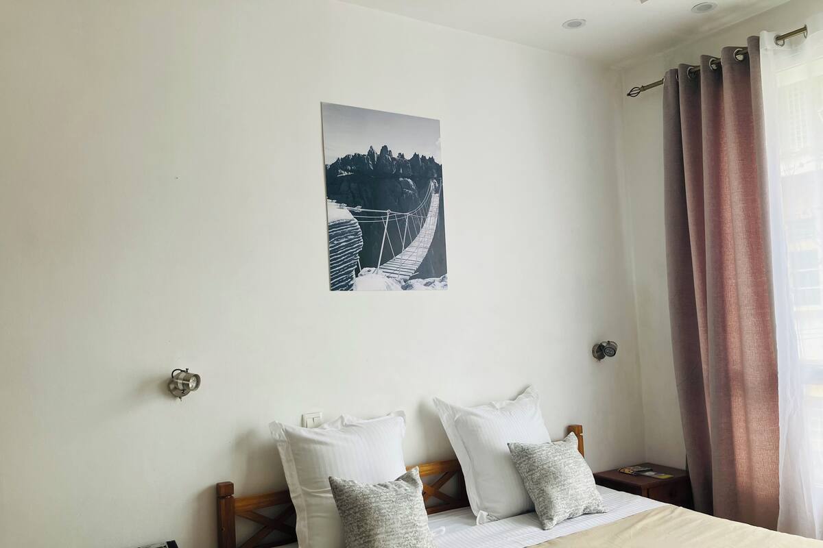 A bedroom with a bed, pillows, a nightstand, a telephone, a wall-mounted picture, and a window with curtains.