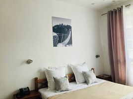 Un dormitorio con cama, almohadas, mesita de noche, teléfono, un cuadro en la pared y ventana con cortinas.