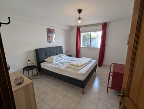 Ein Schlafzimmer mit einem Bett, einem Nachttisch, einer Hängeleuchte, einem Fenster mit Vorhängen und einer Tür.