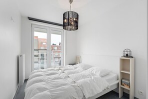 Ein modernes Schlafzimmer mit Bett, Heizkörper, Bücherregal und Blick auf Gebäude durch ein Fenster.
