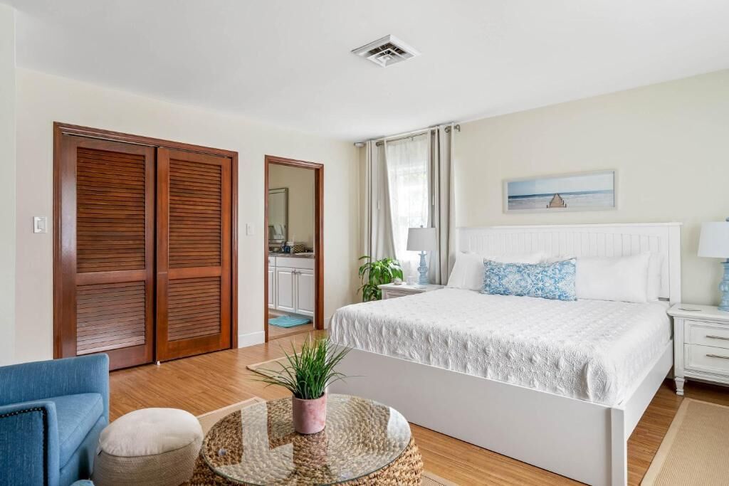 Un dormitorio con cama, una silla, una mesita redonda con una planta, una mesita de noche y una ventana con cortinas.