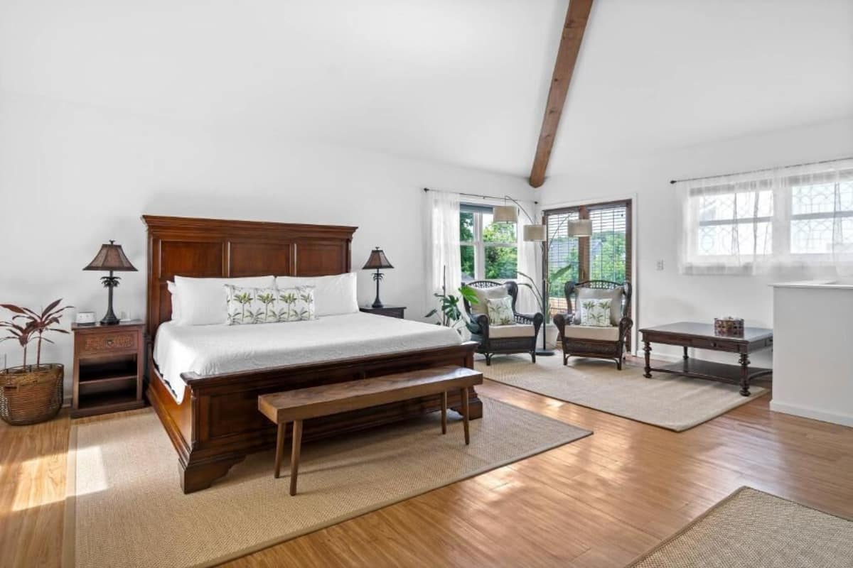 A bedroom with a wooden bed, a bench, a chair, a small table, and a window with curtains.