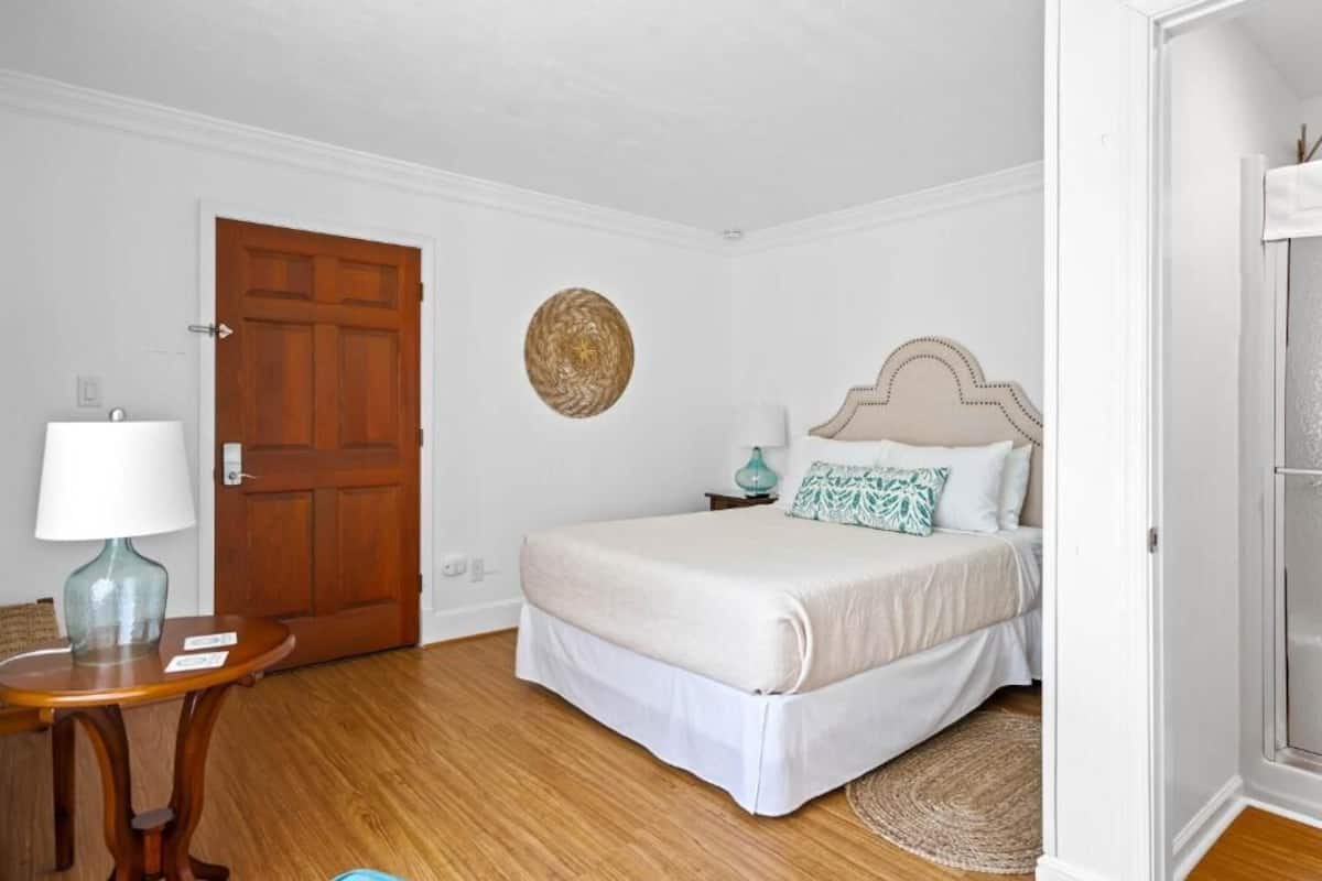 A bedroom with a wooden door, a round woven wall hanging, a bed with white bedding, a small table with a lamp, and a woven rug.
