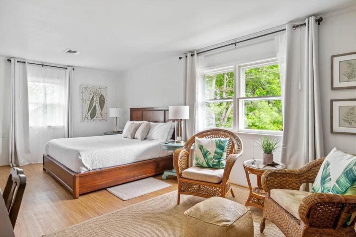 A bedroom with a large bed, a chair, a small table, and a window with a view of greenery.