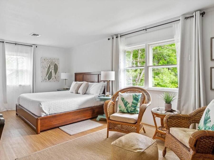 A bedroom with a large bed, a chair, a small table, and a window with a view of greenery.