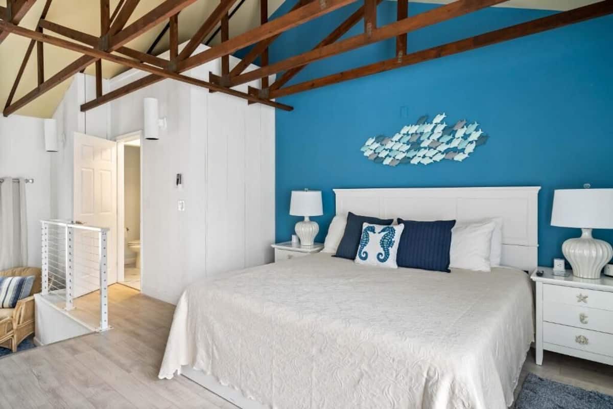 A bedroom with a large bed, white nightstands, a blue accent wall, and exposed wooden beams.