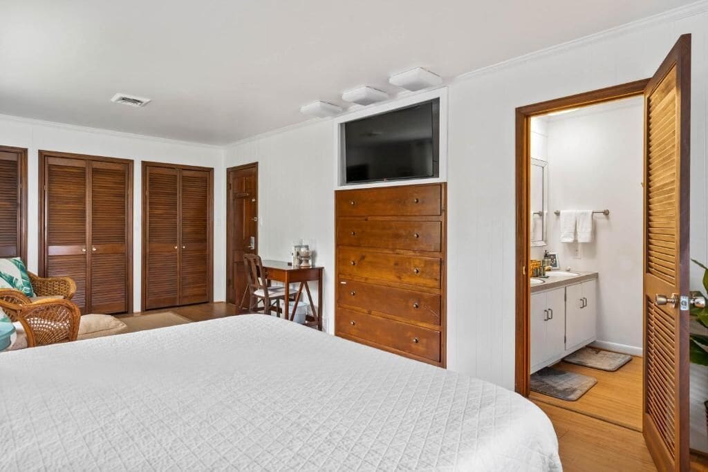 A bedroom with a large bed, a wooden dresser, a desk with a chair, and a bathroom with a mirror and sink.