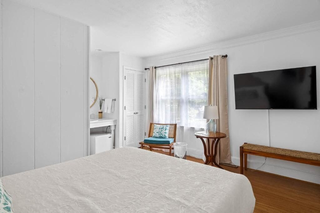 Habitación de hotel con cama, televisión, silla, mesita y ventana con cortinas.