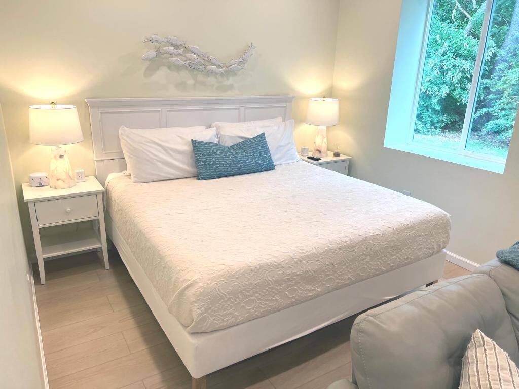 Un dormitorio con una cama grande, dos mesitas de noche con lámparas, un adorno en la pared y una ventana con vistas a la vegetación.